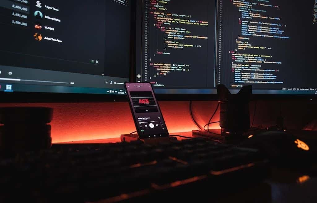A dark desk lit by warm light, two monitors showing code, and a phone running a retro NES app.