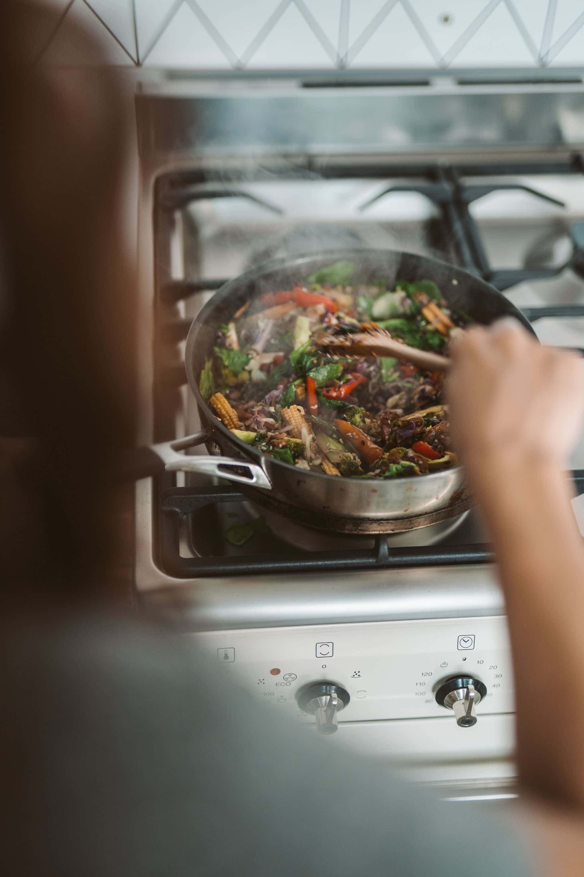 A person cooking a colorful vegetable stir-fry in a pan on a gas stove.