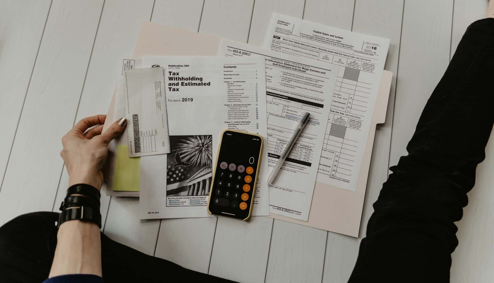 A person sitting on the floor with tax forms, an envelope, a pen, and a phone calculator.