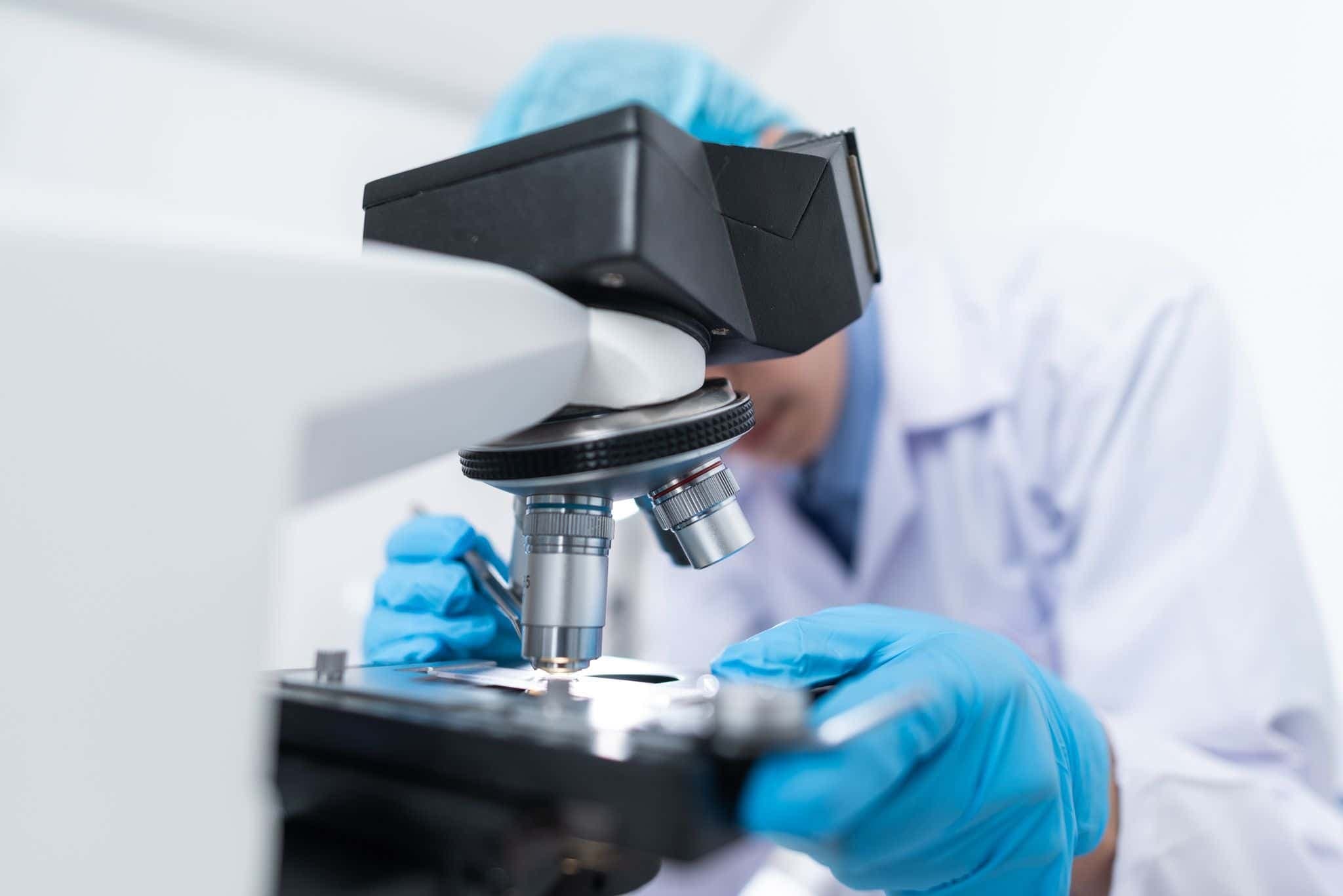A gloved scientist adjusting a slide under a microscope in a bright lab.