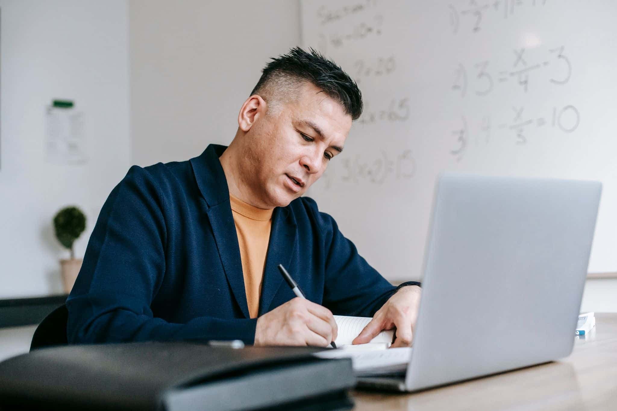 A researcher writing notes beside a laptop, equations on the whiteboard behind him.