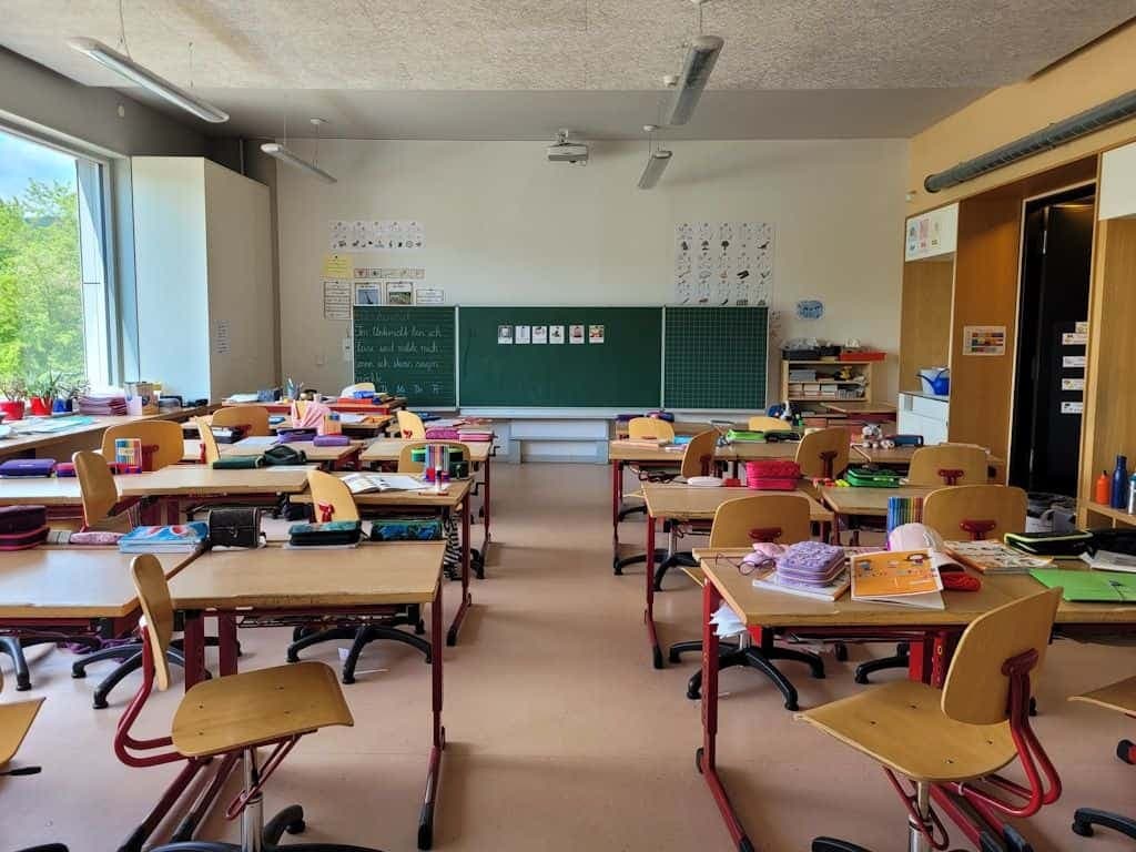 An empty classroom with wooden desks, supplies laid out, and a green chalkboard at the front.