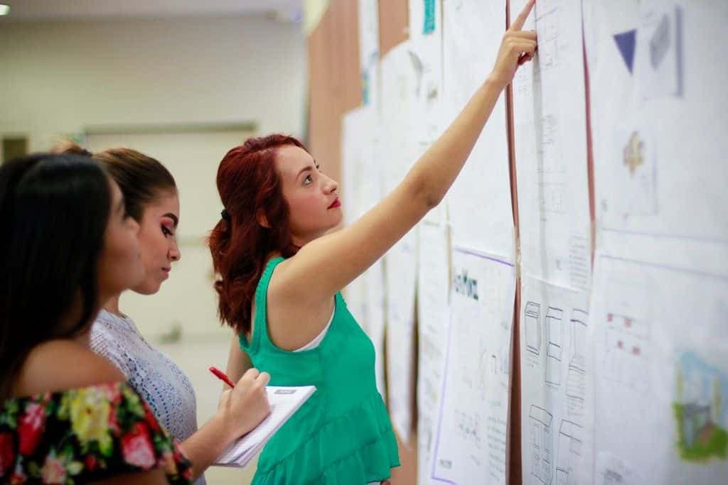 A teacher points at work pinned to a wall while two students take notes beside her.