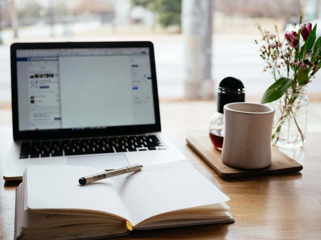 An open notebook and pen in front of a laptop, with coffee and flowers on a wooden desk by a window.