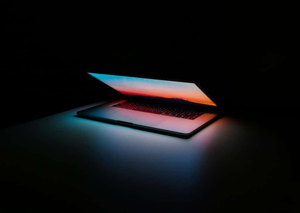 A MacBook opening in a dark room, its screen casting colored light across the keyboard.