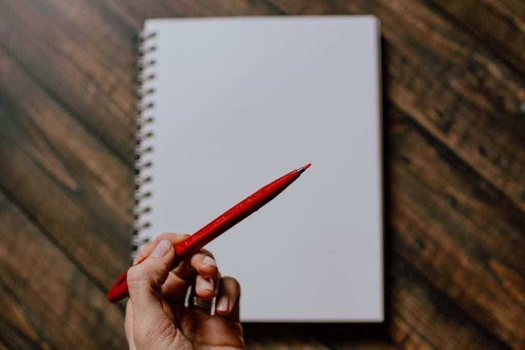 A hand holding a red pen over a blank spiral notebook on a wooden desk.