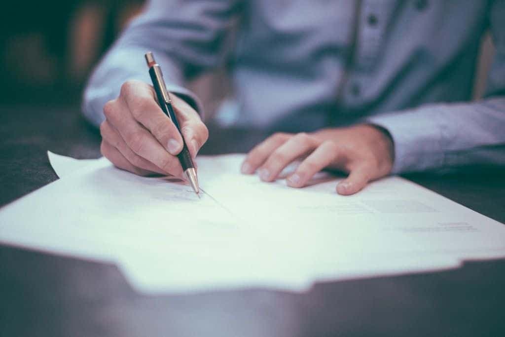 A person in a blue shirt signing a stack of paper documents with a pen.