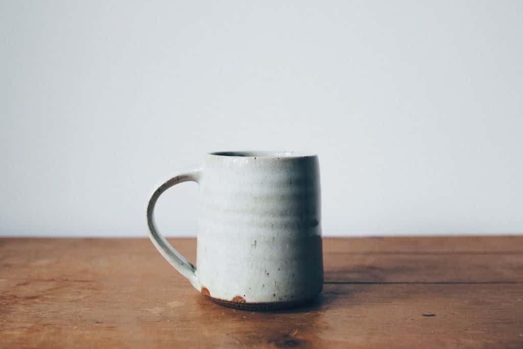 A handmade white ceramic mug sitting on a warm wooden tabletop in soft morning light.