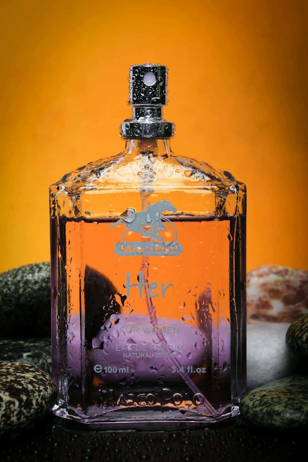 A glass perfume bottle with an etched logo and label on a dark stone surface against a warm backlight.