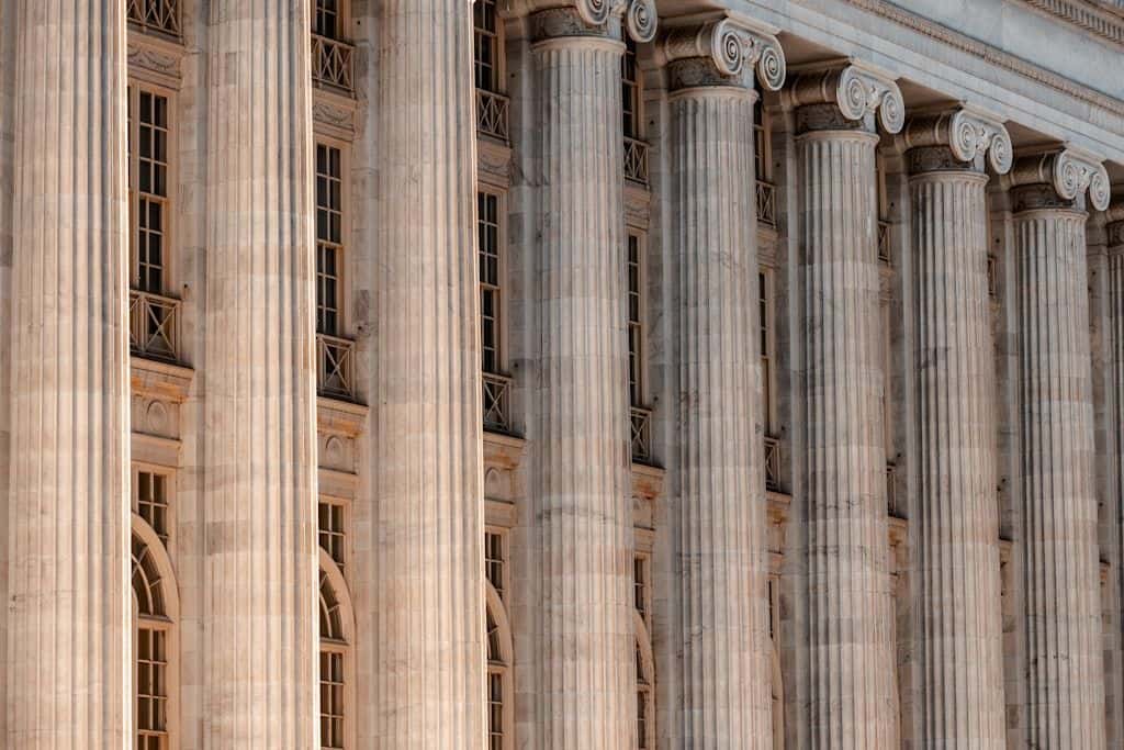 A row of tall classical Ionic columns on a neoclassical government building facade, warm late-afternoon light raking across the marble.
