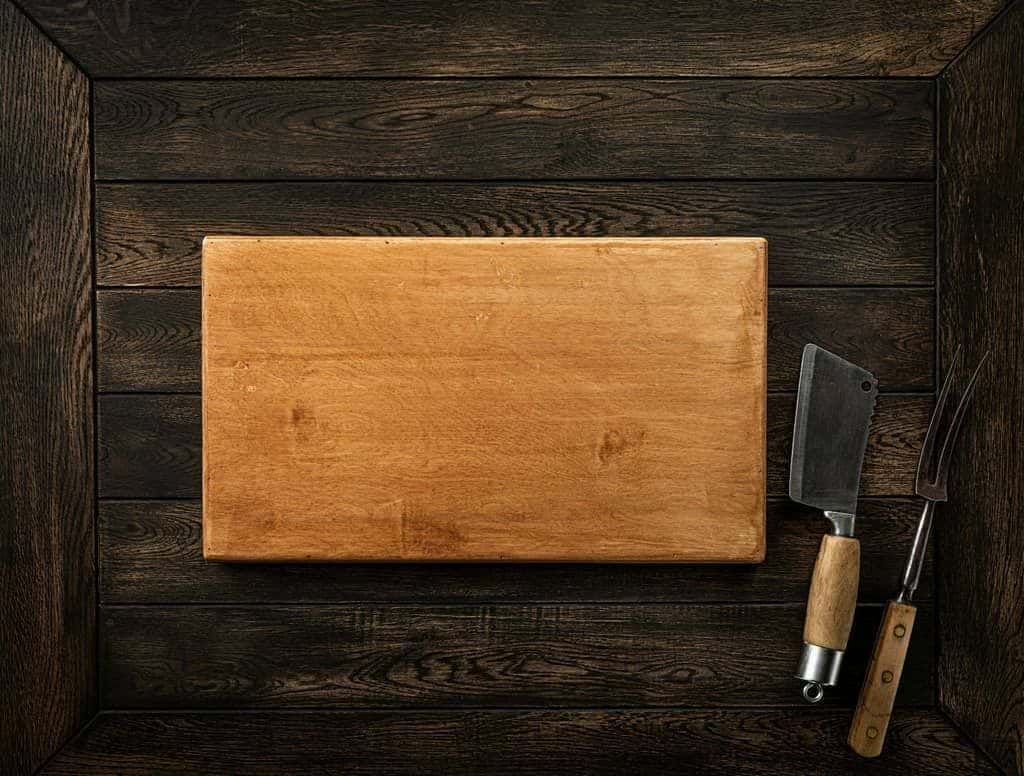 A worn wooden cutting board next to an antique butcher knife and fork on dark wooden planks.