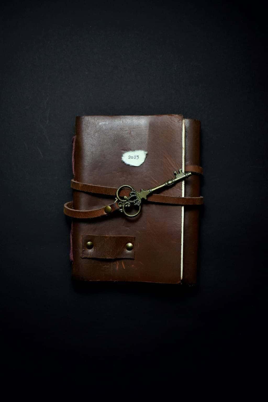 A worn brown leather journal tied shut with a strap and an antique key on a dark background.