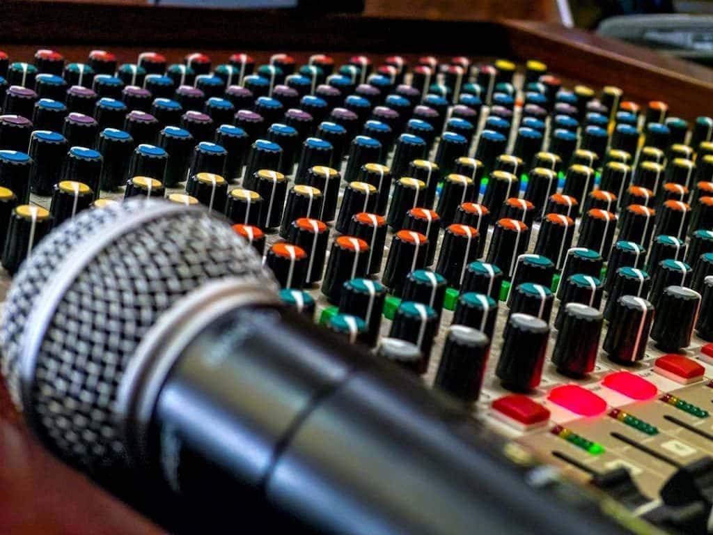 A black and silver studio microphone resting on a live sound mixing board lit by colorful knobs.