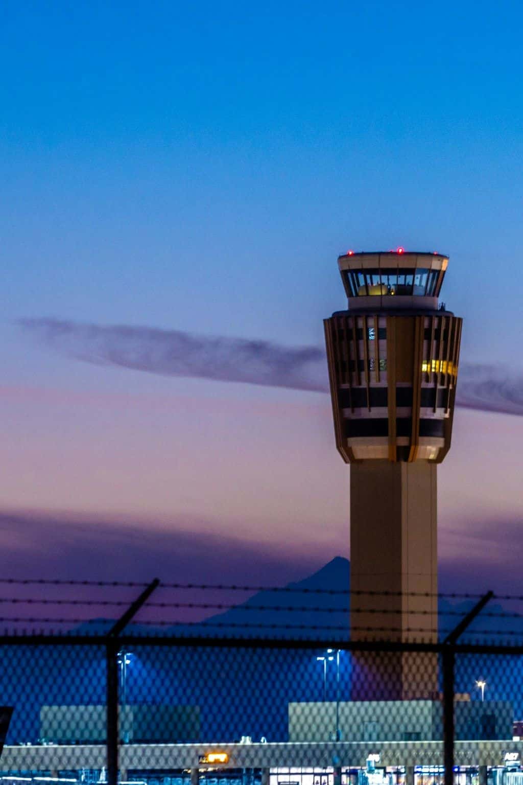 An airport control tower lit at dusk behind a perimeter fence.